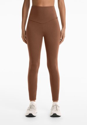 Höga bruna leggings med en slät yta, med en chevrondesign i midjan och sidofickor, parat med ljusa sneakers.