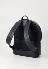 Mochila negra con acabado de cuero texturizado, perfil delgado, respaldo de malla acolchada, correas ajustables y una banda con logotipo en la parte delantera.
