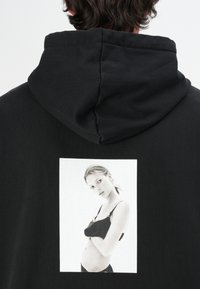 Svart hettegenser med et stort monokromt fototrykk på baksiden, som viser en modell i bh, med en glatt stofftekstur.