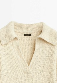 Pull en maille texturée couleur crème avec un large col et un profond décolleté en V, montrant l'étiquette de la marque à l'intérieur du col au dos.