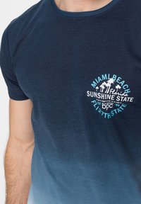 Maglietta in cotone blu navy con design grafico e testo bianco e azzurro chiaro. Collo a giro con un effetto sfumatura di colore.