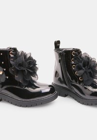 Czarne lakierowane botki za kostkę z ozdobnym czarnym tiulowym kwiatem, złotymi oczkami i zamkiem błyskawicznym z boku dla łatwego zakładania. Gumowa podeszwa.