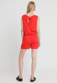 Czerwony kombinezon bez rękawów z wiązaniem z tyłu, elastycznym pasem i szortami. Gładka tkanina z ozdobnymi falbankami na krawędziach. Czarne zdobione baleriny.