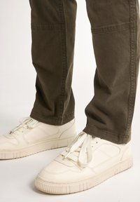 Un pantalon vert olive avec une coupe décontractée et des détails de couture, associé à des baskets blanches avec une surface texturée et des lacets plats.