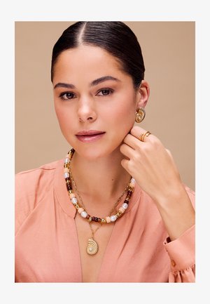 Biżuteria w złotym kolorze z zawieszką w kształcie muszli, kolorowymi koralikami oraz łańcuszkiem, noszona z dopasowanymi kolczykami i pierścionkami.