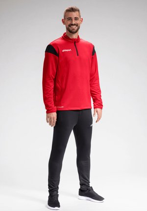 Man staand, draagt een rood-zwarte sporttrui, zwarte sportbroek en zwarte sneakers, glimlacht tegen een effen achtergrond.