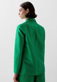 Green blazer with a textured finish, featuring a scalloped collar and long sleeves. The back shows clean lines and structured tailoring.