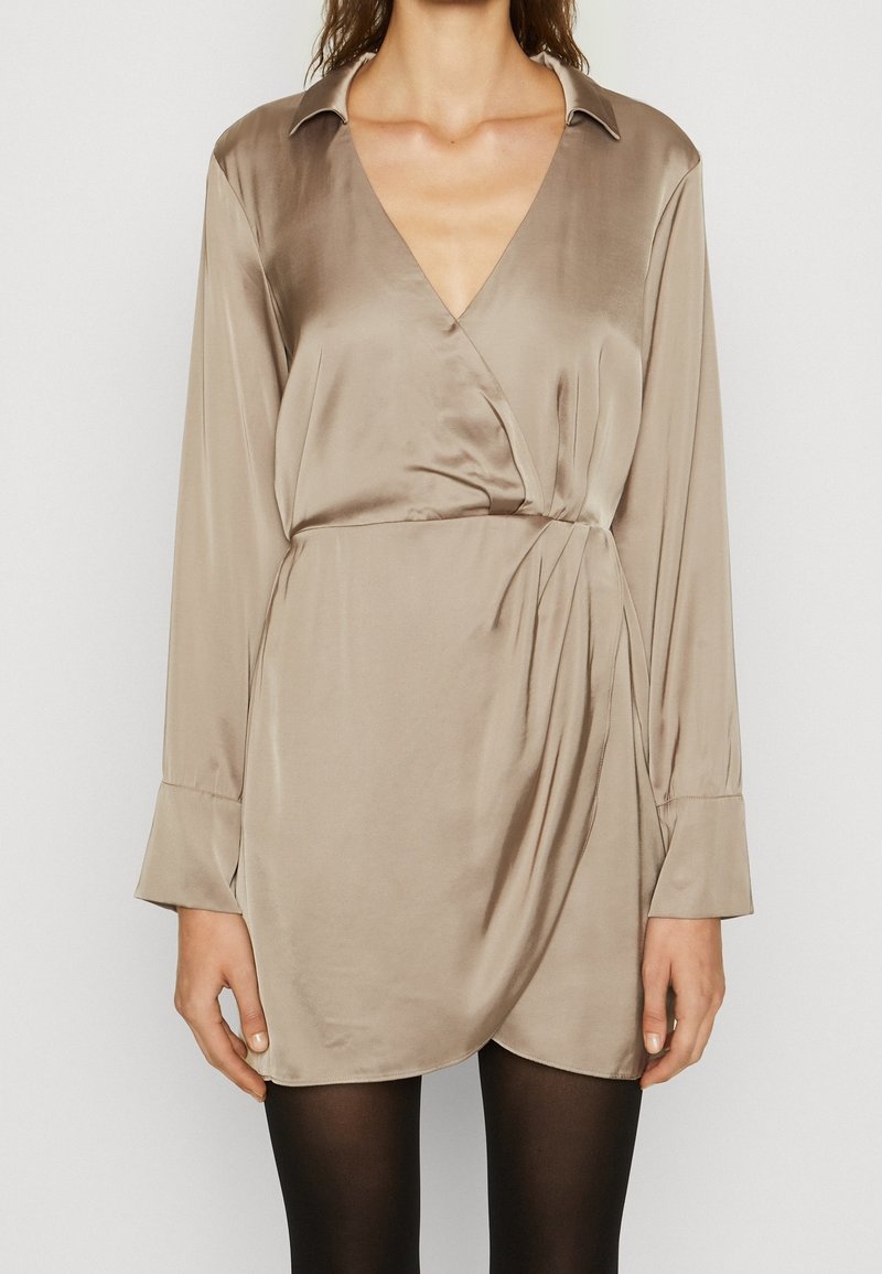 Vestido envolvente de satén beige con mangas largas, escote en V profundo y cintura ajustada. Presenta una textura suave y un brillo sutil.