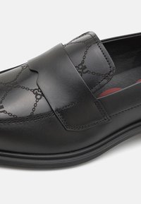 Mocassin en cuir noir à surface texturée, avec un design à bride et un motif de logo subtil. Semelle en caoutchouc robuste et finition lisse.