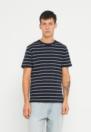 Tricou cu dungi marine, cu dungi orizontale albe, guler rotund, mâneci scurte, din material de bumbac, purtat cu blugi de culoare albastru deschis.