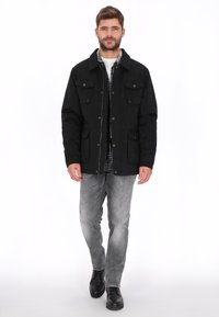 Zwarte jas met een kraag, knoopsluiting aan de voorkant en vier opgestikte zakken. Draagt deze over een geruit overhemd, gecombineerd met lichtgrijze spijkerbroek en zwarte laarzen.