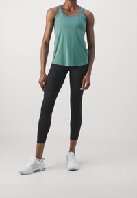 Vrouw staat met een teal mouwloos sporttop, zwarte strakke leggings en witte hardloopschoenen tegen een effen lichte achtergrond.