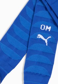 Blauwe sokken met een gestructureerd golfpatroon, voorzien van een contrasterend wit Puma-logo en "OM" letters. Gemaakt van rekbaar breiwerk.