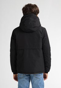 Giacca nera con cappuccio e texture liscia, dotata di chiusura con zip e polsini elasticizzati. Indossata con jeans azzurri, vista da dietro.