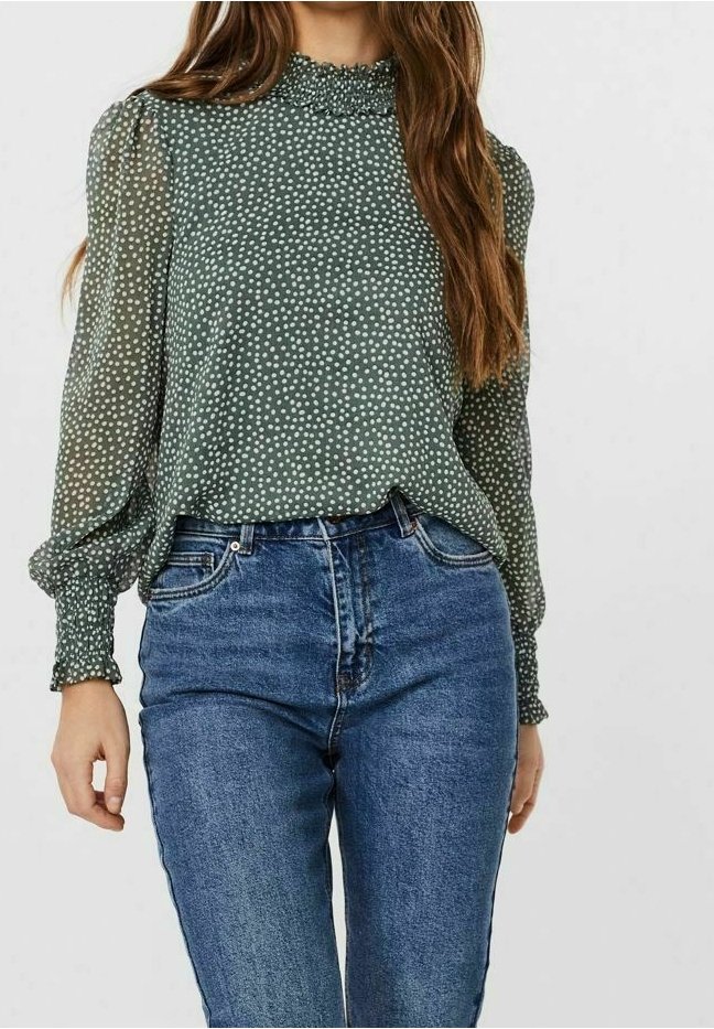 Blouse verte transparente à manches longues avec des pois blancs rentrée dans un jean en denim bleu taille haute portée par une femme.