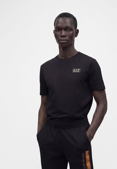 Мъж, облечен в черна тениска Emporio Armani EA7 и черен панталон с оранжев лого, стои с ръце в джобовете, неутрален фон.