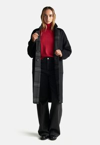 Schwarzer langer Mantel mit grauen Karos, kombiniert mit einem roten Rollkragenpullover und schwarzen hochgeschnittenen Hosen. Glattes Material und lässige Passform.