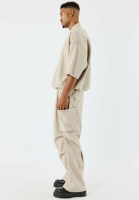 Chemise beige à manches courtes avec une coupe décontractée, associée à un pantalon ample assorti avec une poche à rabat et des chaussures à plateforme noires.