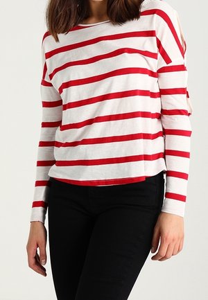 Kobieta ubrana w białą koszulę z długim rękawem z poziomymi czerwonymi paskami i czarne spodnie, stojąca na jednolitej, jasnej ścianie.