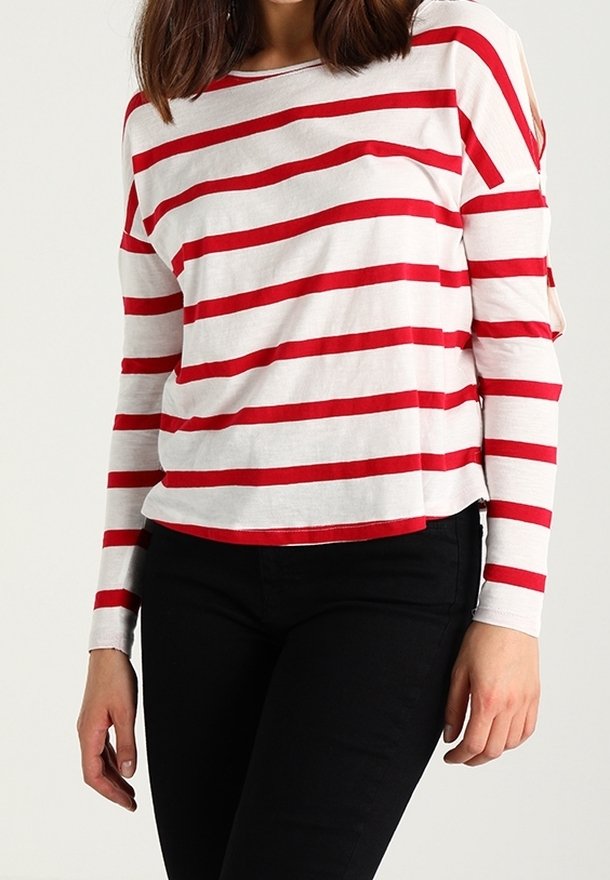 Femme portant une chemise blanche à manches longues avec des rayures rouges horizontales et un pantalon noir, debout devant un fond uni.