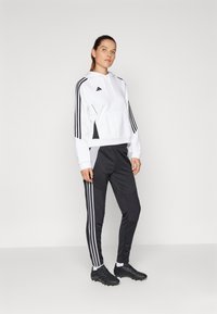 White hooded sweatshirt with black accents, featuring three black stripes on sleeves. Paired with black athletic pants and black cleats.
