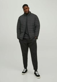 Sort polstret jakke med høj krave, båret over en mørk sweater, parret med sorte tætsiddende bukser og sorte sneakers. Glat tekstur, minimalistisk design.
