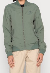 Zielona kurtka bomberka z zapięciem na zamek, prążkowanym kołnierzem i mankietami, kieszeniami po bokach oraz gładką fakturą. Ukryty detal z logo na klatce piersiowej.