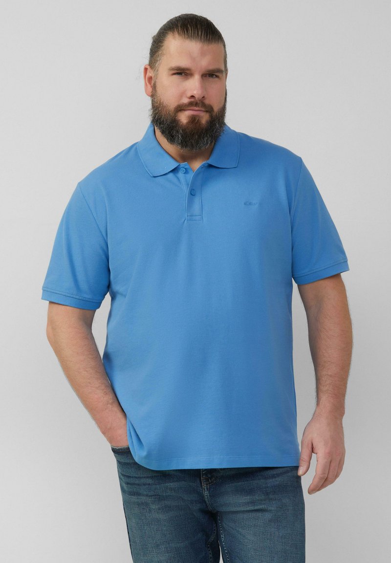 Lichtblauw poloshirt met een kraag, korte mouwen en een subtiele logo op de linkerborst. Draagt met donkere jeans, textuur zichtbaar.