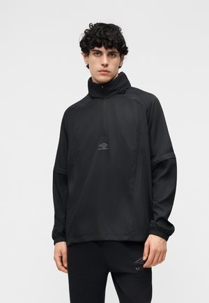 Mladý muž s kučeravými vlasmi, oblečený v čiernej pulóvrovej mikine Umbro s vysokým golierom a v zodpovedajúcich čiernych nohaviciach, stojaci pred jednoduchým svetlým pozadím.