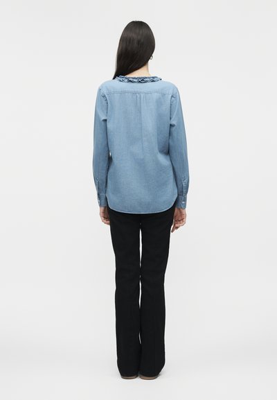 Γυναίκα με μακριά σκουρόχρωμα μαλλιά που φοράει ένα τζιν πουκάμισο με ανοιχτό μπλε ρεβέρ με βολάν και μαύρο παντελόνι, στέκεται με την πλάτη προς την κάμερα σε απλό φόντο.