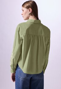 Camicia verde chiaro con bottoni, dotata di una piega posteriore e decorazioni ricamate sulle maniche, abbinata a jeans blu. Tessuto morbido, vestibilità casual.