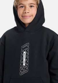 Čierna mikina s kapucňou s veľkým bielym logom na prednej strane. Materiál vyzerá mäkko s uvoľneným strihom a priestrannou kapucňou.