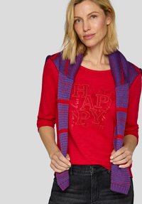 Rotes langärmliges Hemd mit dekorativem Text, kombiniert mit einem blau-lila gemusterten Schal, der über die Schultern drapiert ist. Dunkle Jeans.