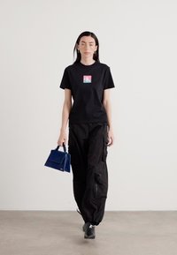 Black t-shirt with colorful logo, cargo pants, and small blue handbag. Model wears athletic shoes and stands on a neutral background.