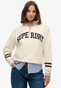 Crème hoodie met rits met "SUPER DRY" in marineblauw, voorzien van zwarte en witte strepen op de mouwen. Gelaagd over een blauw en roze gestreepte shirt.