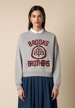 Vrouw met stijl donker haar draagt een grijze trui met de tekst en het logo van "Brooks Brothers" in het rood, gecombineerd met een geplooide donkerblauwe rok.
