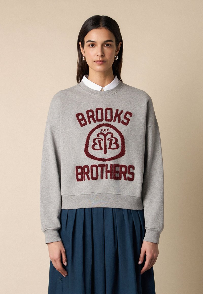 Vrouw met stijl donker haar draagt een grijze trui met de tekst en het logo van "Brooks Brothers" in het rood, gecombineerd met een geplooide donkerblauwe rok.
