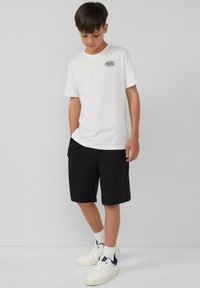 Witte katoenen t-shirt met zwart "PARADISE" logo, gecombineerd met zwarte shorts en witte sneakers met blauwe accenten. Glad materiaal, casual pasvorm.