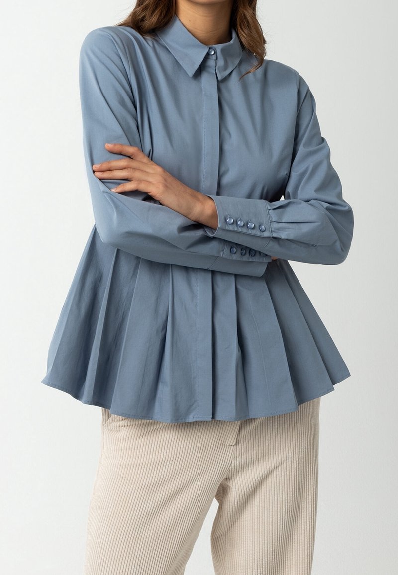 Blusa azzurro chiaro a maniche lunghe con orlo plissettato, polsini con bottoni e colletto. Abbinata a pantaloni in corduroy beige.