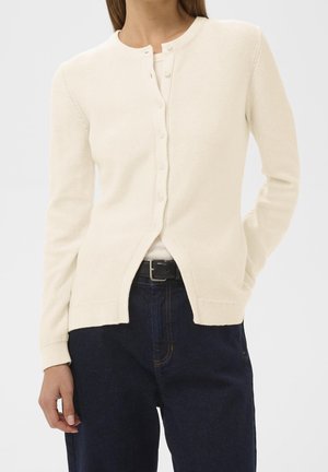 Vrouw draagt een crème-gekleurde gebreide cardigan met knopen over een witte top, donkerblauwe spijkerbroek met een zwarte riem, één hand in de zak, tegen een effen achtergrond.