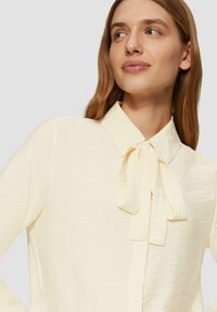Crème-kleurige blouse met een textuurafwerking. Bevat een kraag en een gestrikt strikdetail bij de hals. Lange mouwen en een relaxte pasvorm.