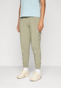 Persoon in een lichtblauw shirt, olijfgroene broek, witte sokken en beige sportschoenen die op een effen achtergrond staat.