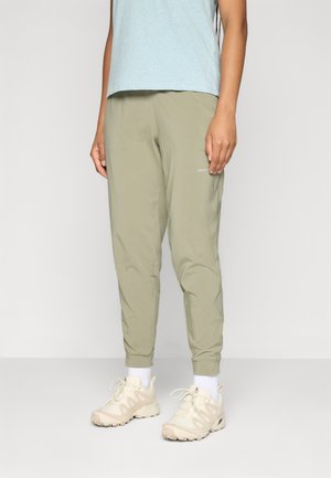 Person som har på seg en lys blå skjorte, olivengrønne bukser, hvite sokker og beige joggesko, stående på en ensfarget bakgrunn.