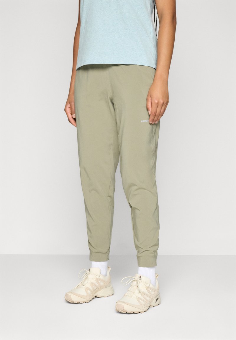 Persoon in een lichtblauw shirt, olijfgroene broek, witte sokken en beige sportschoenen die op een effen achtergrond staat.