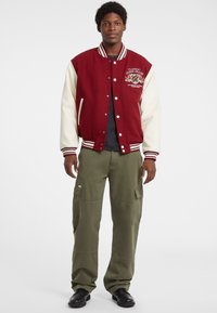 Burgundy ja tõmmise värvi varsity jakk ribaga kaeluse, eesnuppude ja broderitud logoga; koos oliivroheliste cargo-pükste ja mustade kingadega.