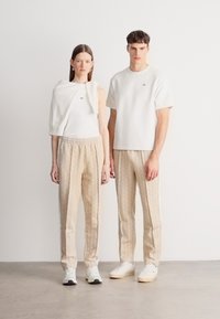 Λευκό κοντομάνικο πουκάμισο και κρεμ φόρμες με διακριτικό σχέδιο για τον άντρα· λευκή φανέλα και κρεμ παντελόνι με σχέδιο για τη γυναίκα.