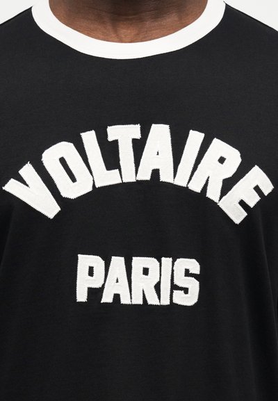 Κλείσιμο σε μαύρο πουκάμισο με λευκό γιακά και τις λέξεις "VOLTAIRE" κυρτές πάνω από το "PARIS" σε μεγάλα λευκά κεντημένα γράμματα στο στήθος.