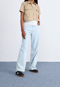 Veste cropped beige avec poches avant, top blanc à côtes, jean large bleu clair et baskets noires avec semelles en gomme. Design simple.