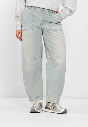 Personne portant un jean large délavé clair avec de grandes poches avant, associé à des baskets New Balance argentées et grises.