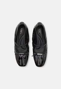 Black patent leather flats with a rounded toe, featuring a small bow accent and a silver DKNY logo on the front. Smooth texture.
